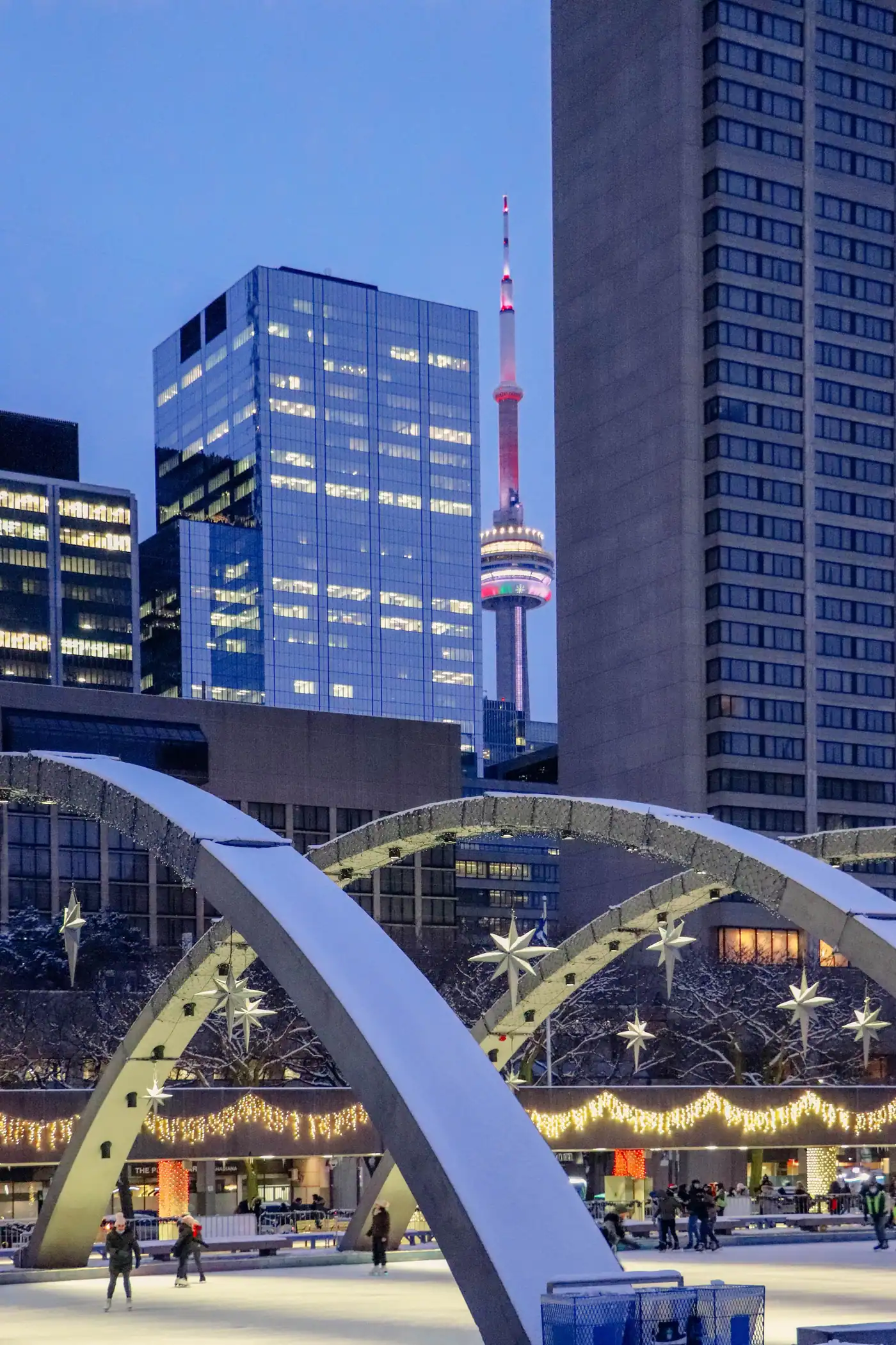 Illuminated arches over a winter square at dusk, the CN Tower and glass towers behind