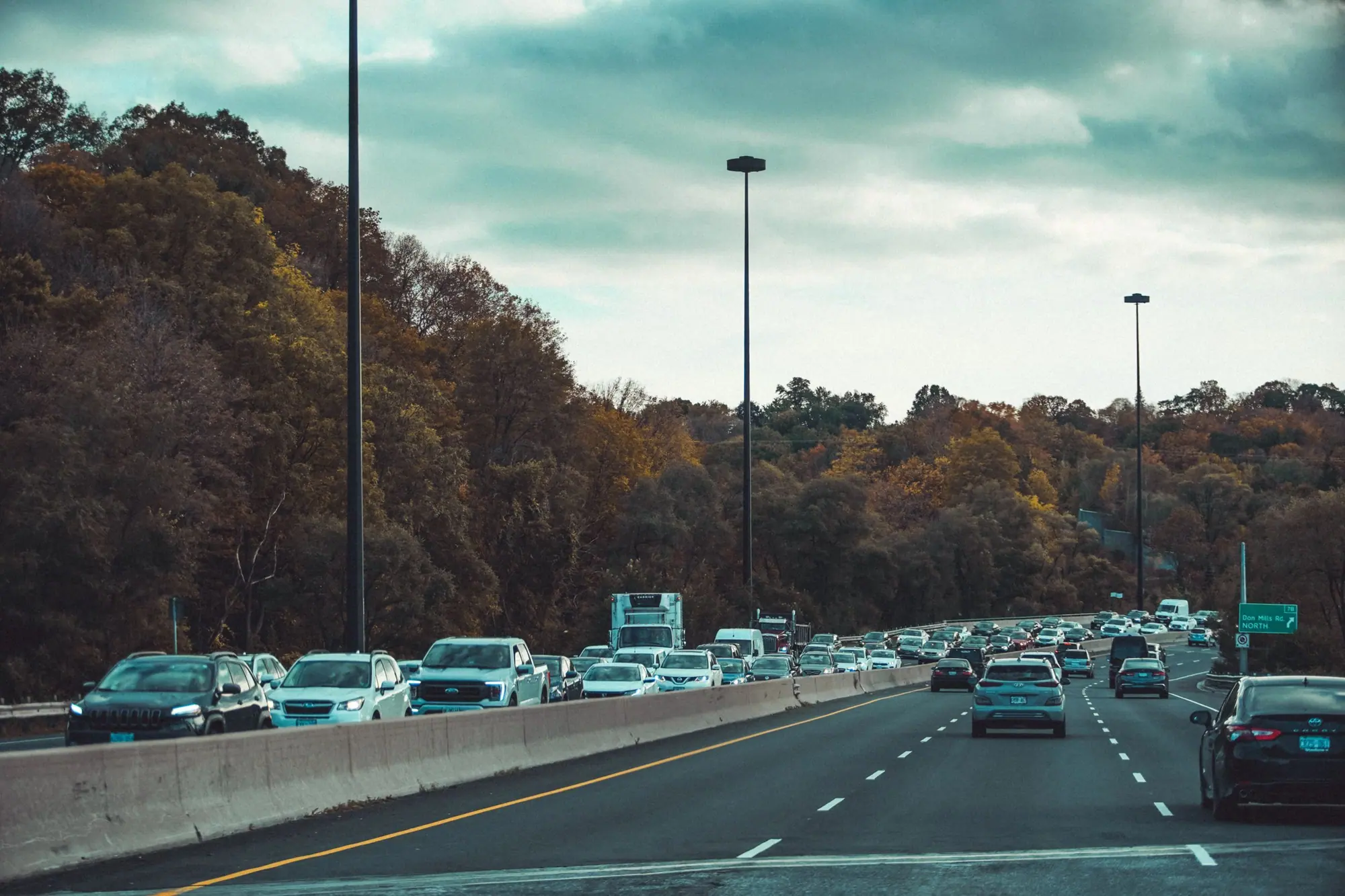 An expressway lined with autumn trees under a heavy teal sky, traffic heading into the city
