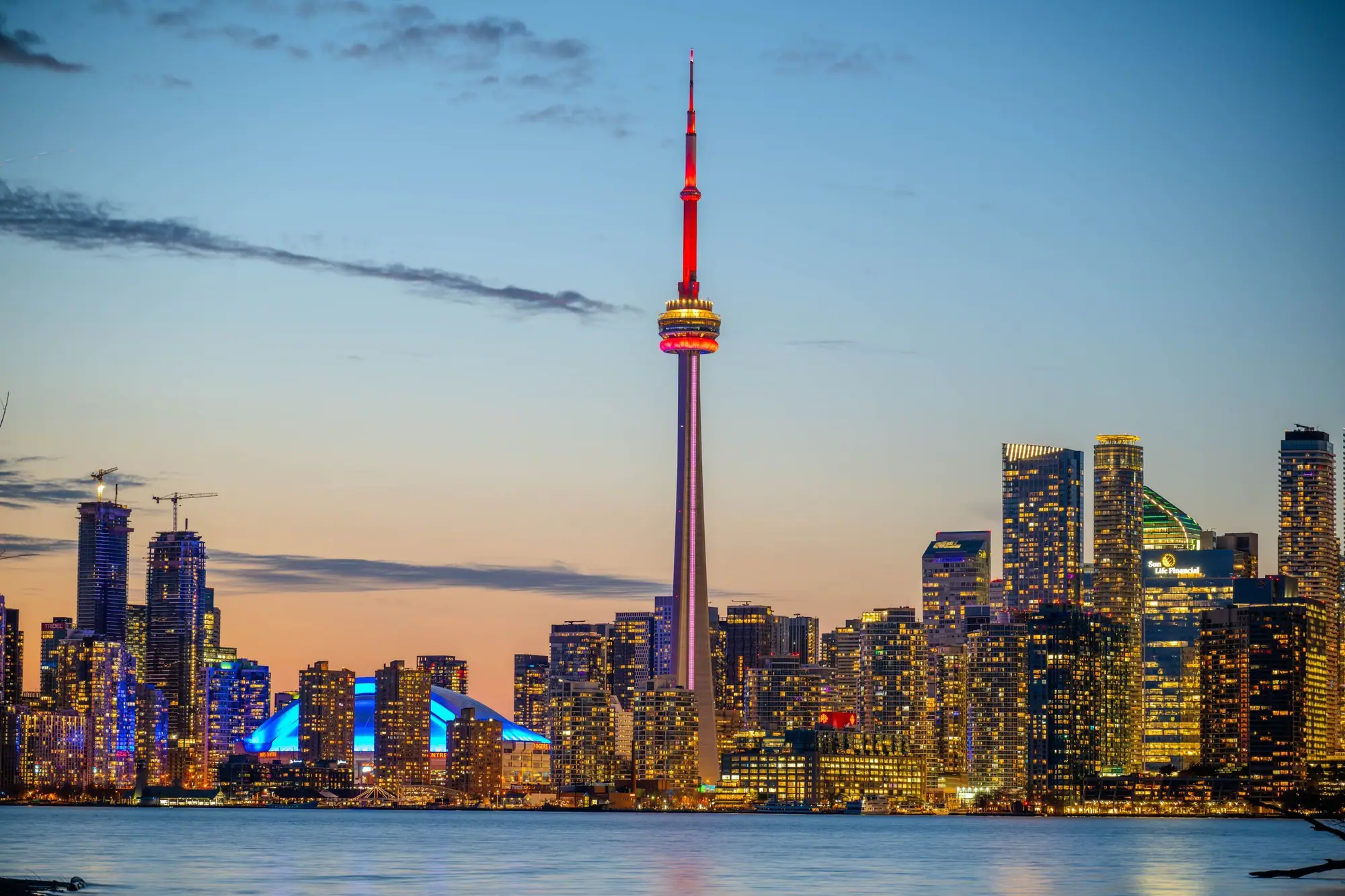 The CN Tower and lit downtown towers beside the lake at sunset, orange light fading into blue sky